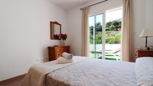 Quarto de hotel com cama, penteadeira, espelho, vaso com flores e vista para o verde pela janela.