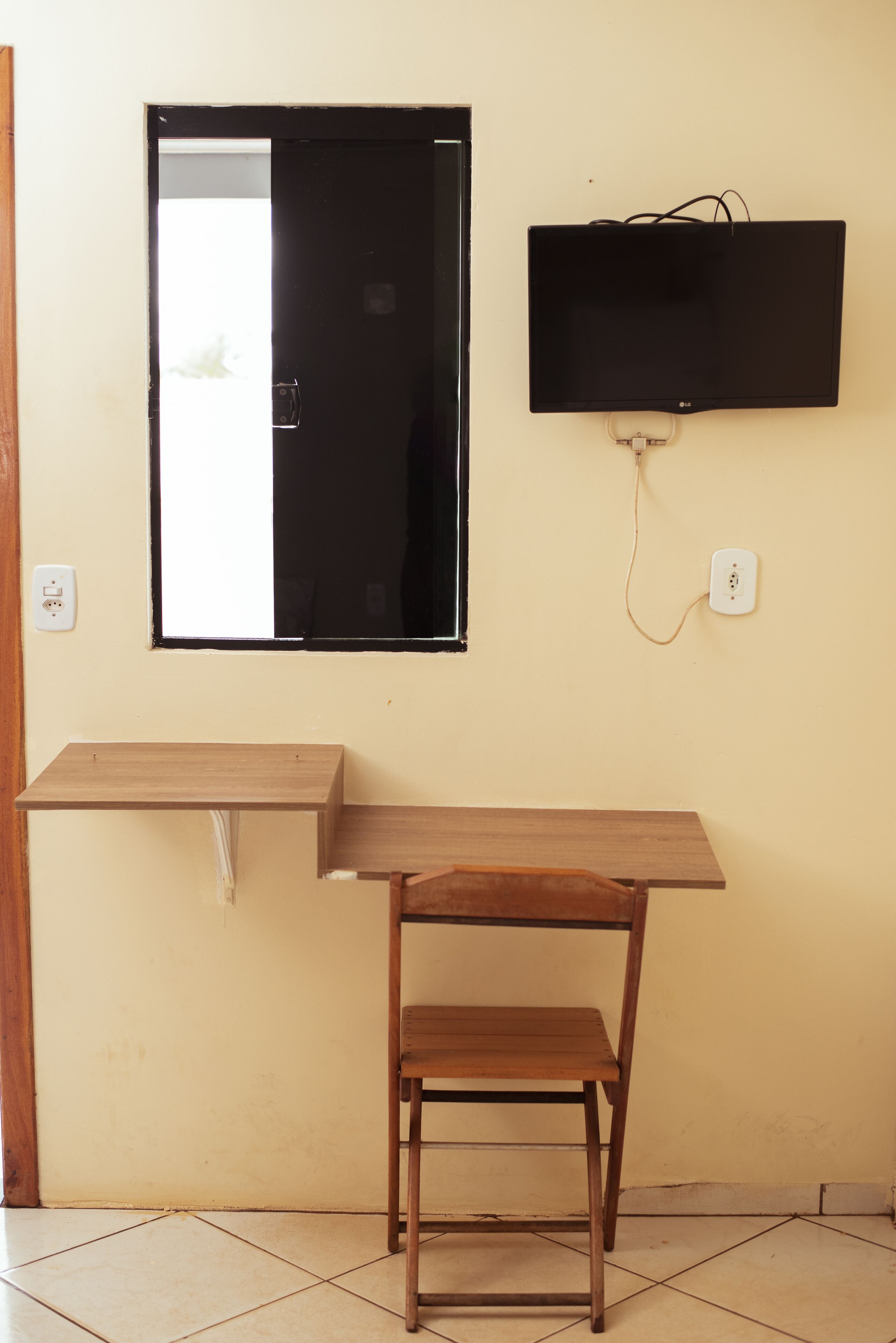 Une pièce comprenant un bureau en bois, une chaise, un téléviseur à écran plat fixé au mur et une fenêtre dotée d’un cadre noir.