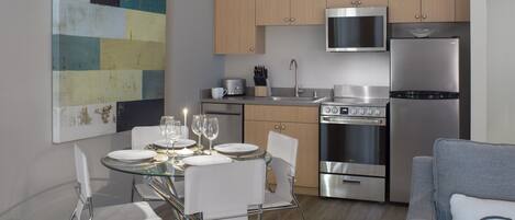 A modern kitchen with stainless steel appliances, a dining area with a glass table, and a grey sofa.