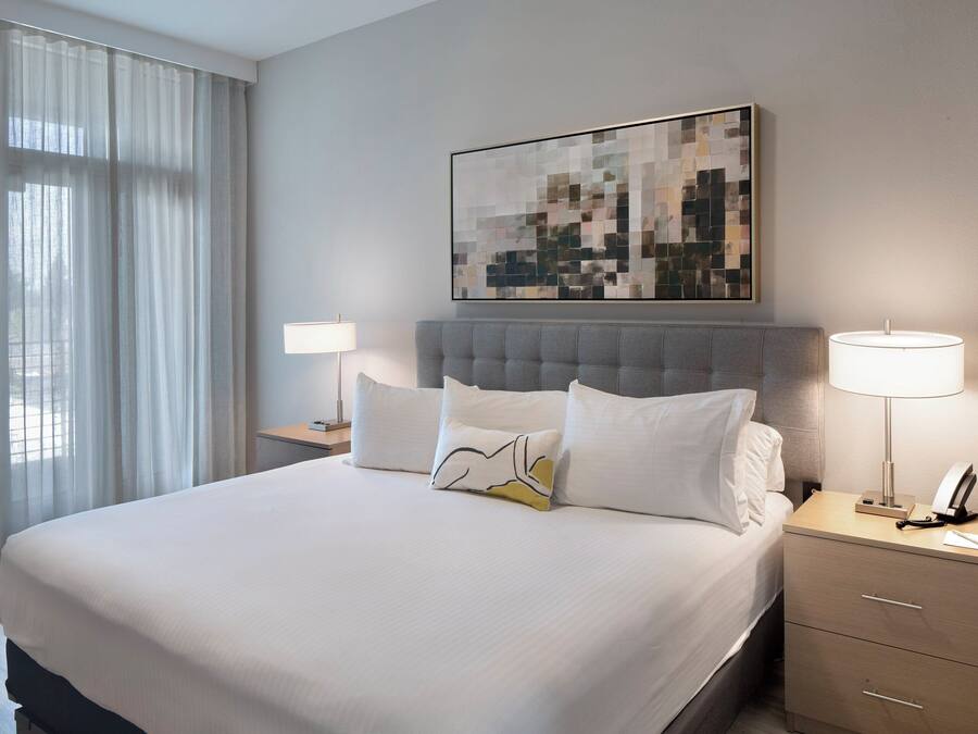 A hotel room with a large bed, a headboard, two bedside tables with lamps, a wall-mounted artwork, and sheer curtains.