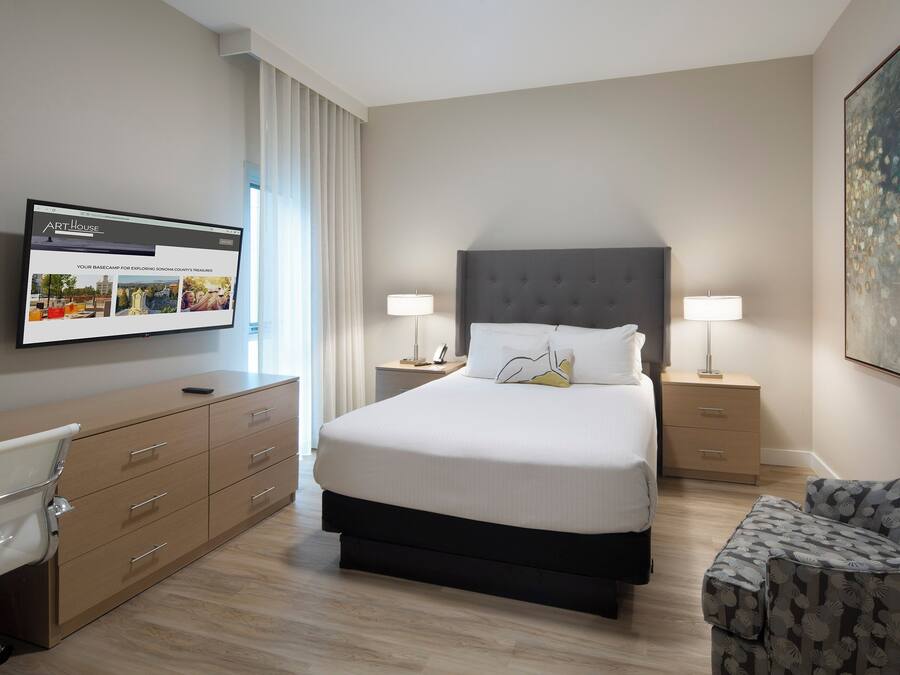A hotel room with a large bed, bedside tables with lamps, a desk with a chair, and a large painting on the wall.