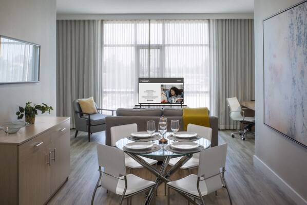 A modern dining area with a glass table set for four, surrounded by white chairs. A grey sofa with a yellow pillow is in the background, and a large window with curtains is visible. - Art House Hotel and Suites (Santa Rosa)