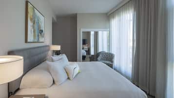 A hotel room with a large bed, a bedside table with a lamp, a chair, and a view of the outside through sheer curtains.