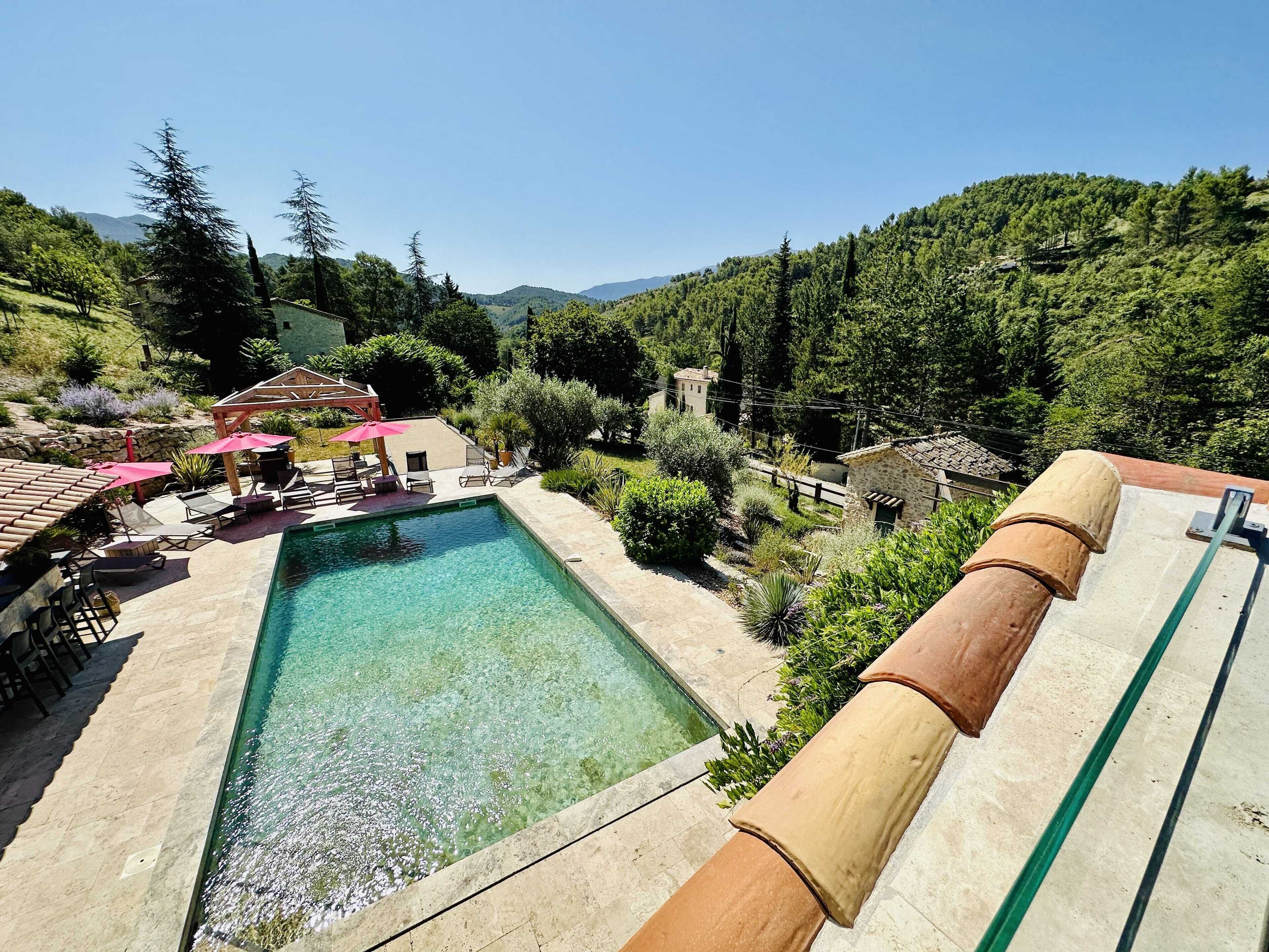 A swimming pool with clear water, surrounded by lounge chairs and umbrellas, set against a backdrop of lush greenery and hills.