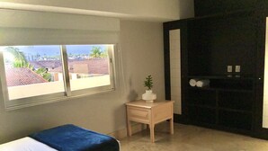 A room with two beds, a small wooden nightstand, a window with a view of buildings and palm trees, and a black door. - Apartments Deck 12 Vallarta (Puerto Vallarta)