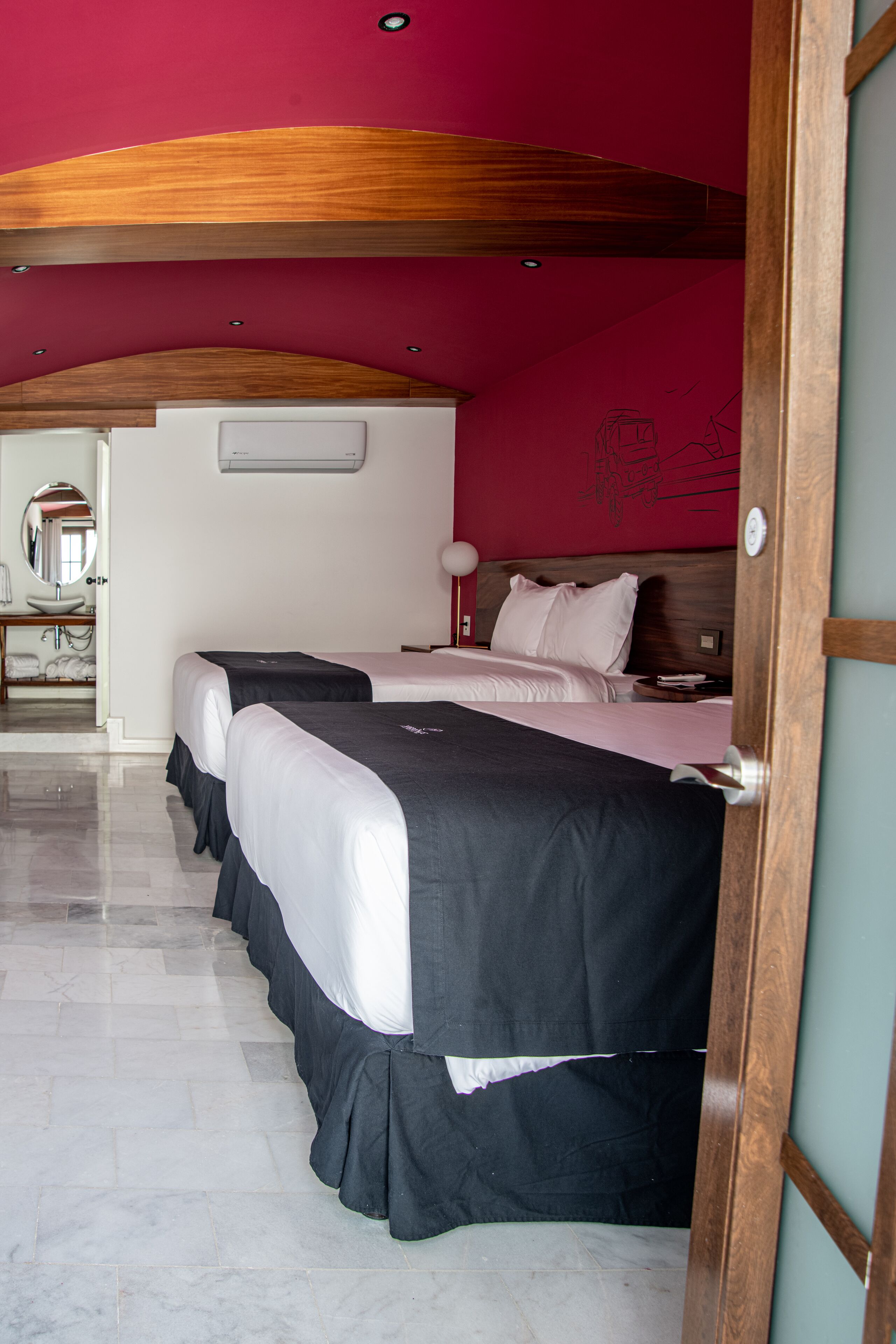 Habitación de hotel con dos camas, techo de madera, un espejo y un baño visible a través de una puerta abierta.