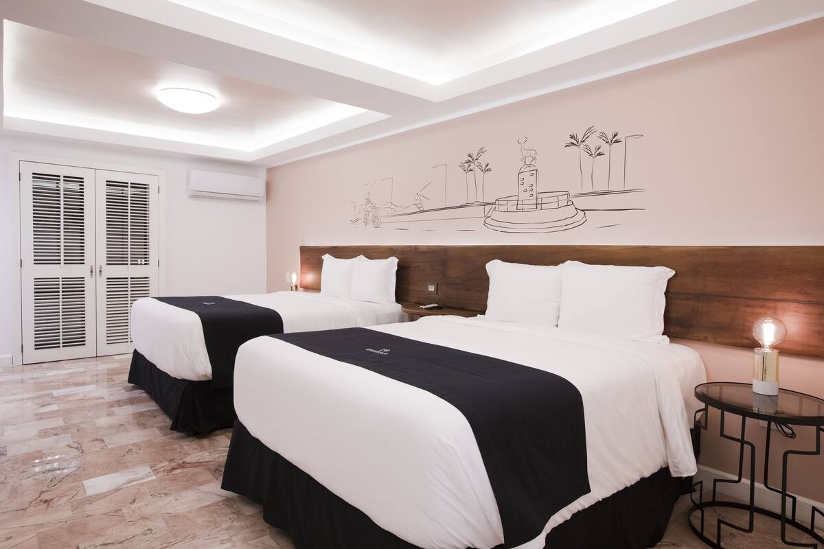 Habitación de hotel con dos camas, un cabecero de madera, un mural en la pared, una mesita con lámpara e iluminación en el techo.