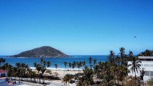 Vista de playa con palmeras, una montaña al fondo y un cielo azul despejado.