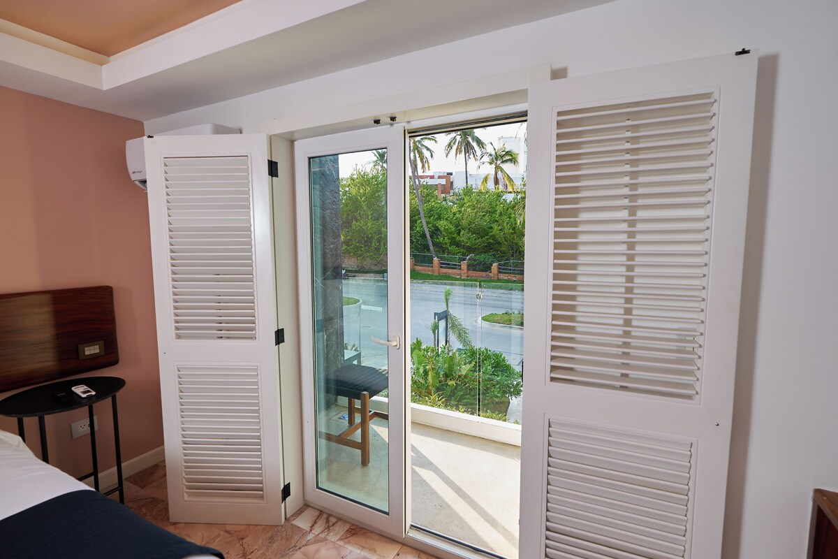 Ein Hotelzimmer mit einem Bett, einem kleinen Tisch, einem Sessel und einer Glasschiebetür, die auf einen Balkon mit Blick auf einen Pool und Grünflächen führt.