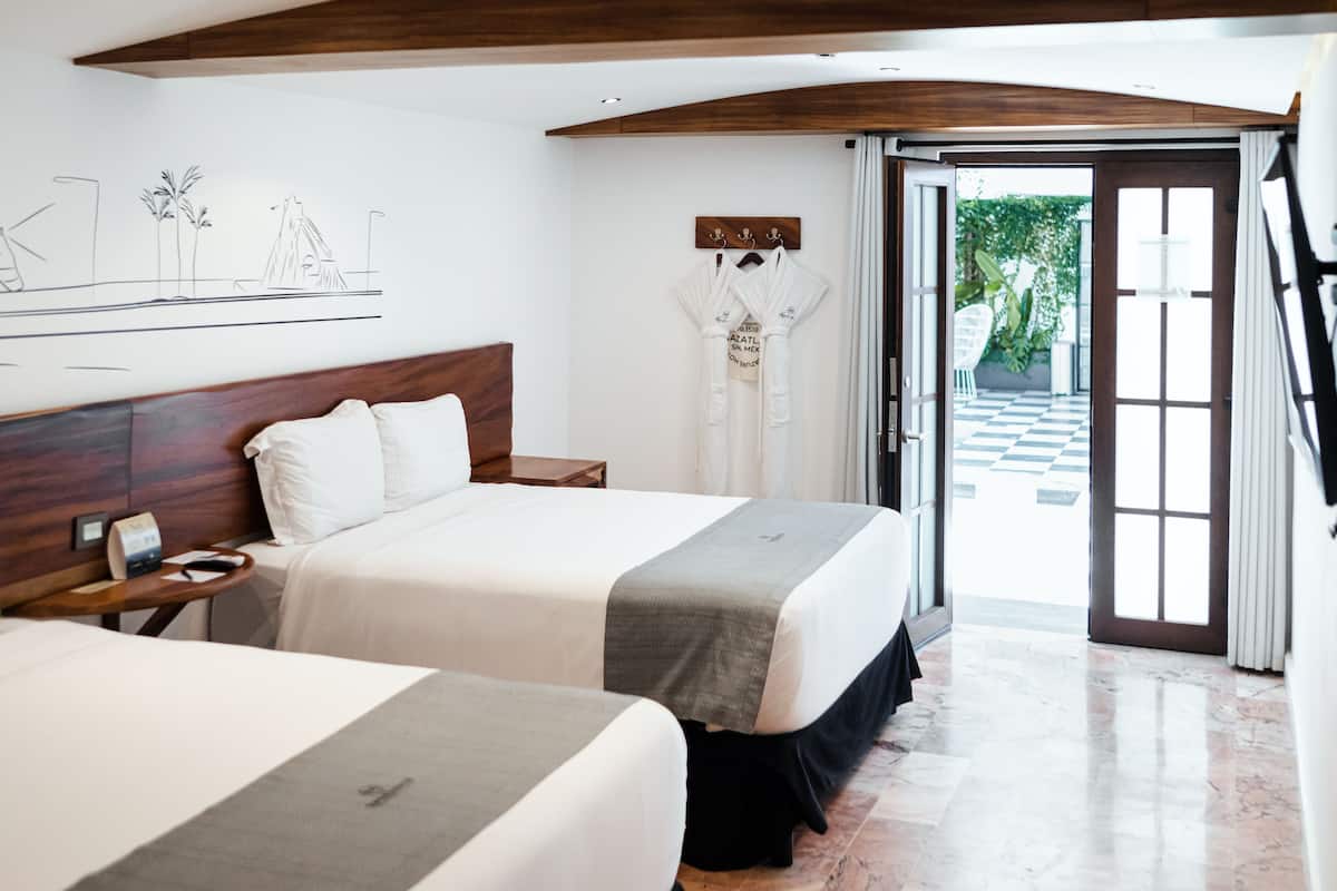 Habitación de hotel con dos camas, cabecera de madera, mesita de noche, un cuadro con un paisaje urbano y vista a un área exterior a cuadros a través de una puerta abierta.