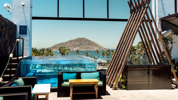 Zona de piscina con cerramiento de vidrio, mobiliario exterior y vista al mar y a la montaña.