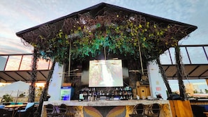 Un bar al aire libre con pared de piedra, taburetes y una pantalla grande.
