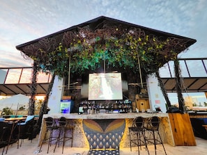 An outdoor bar with a stone wall, bar stools, and a large screen. - The Xperience by g (Mazatlán)