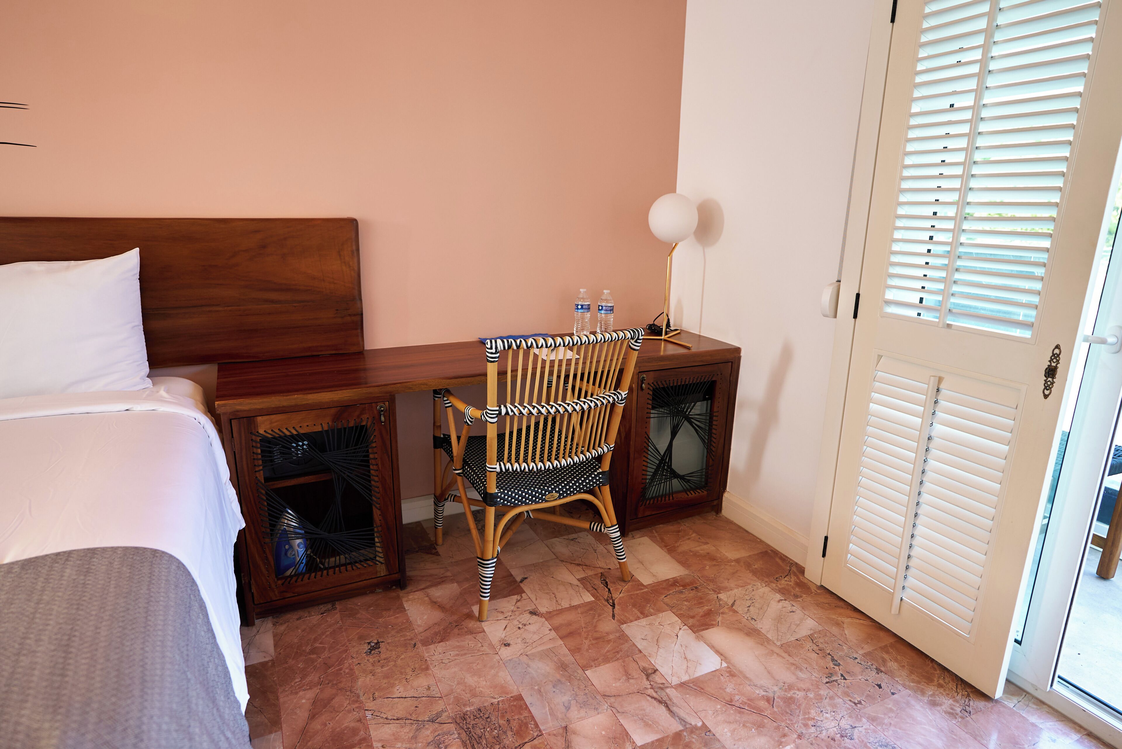 A hotel room with a bed, a desk with two bottles, a chair, and a door with shutters.