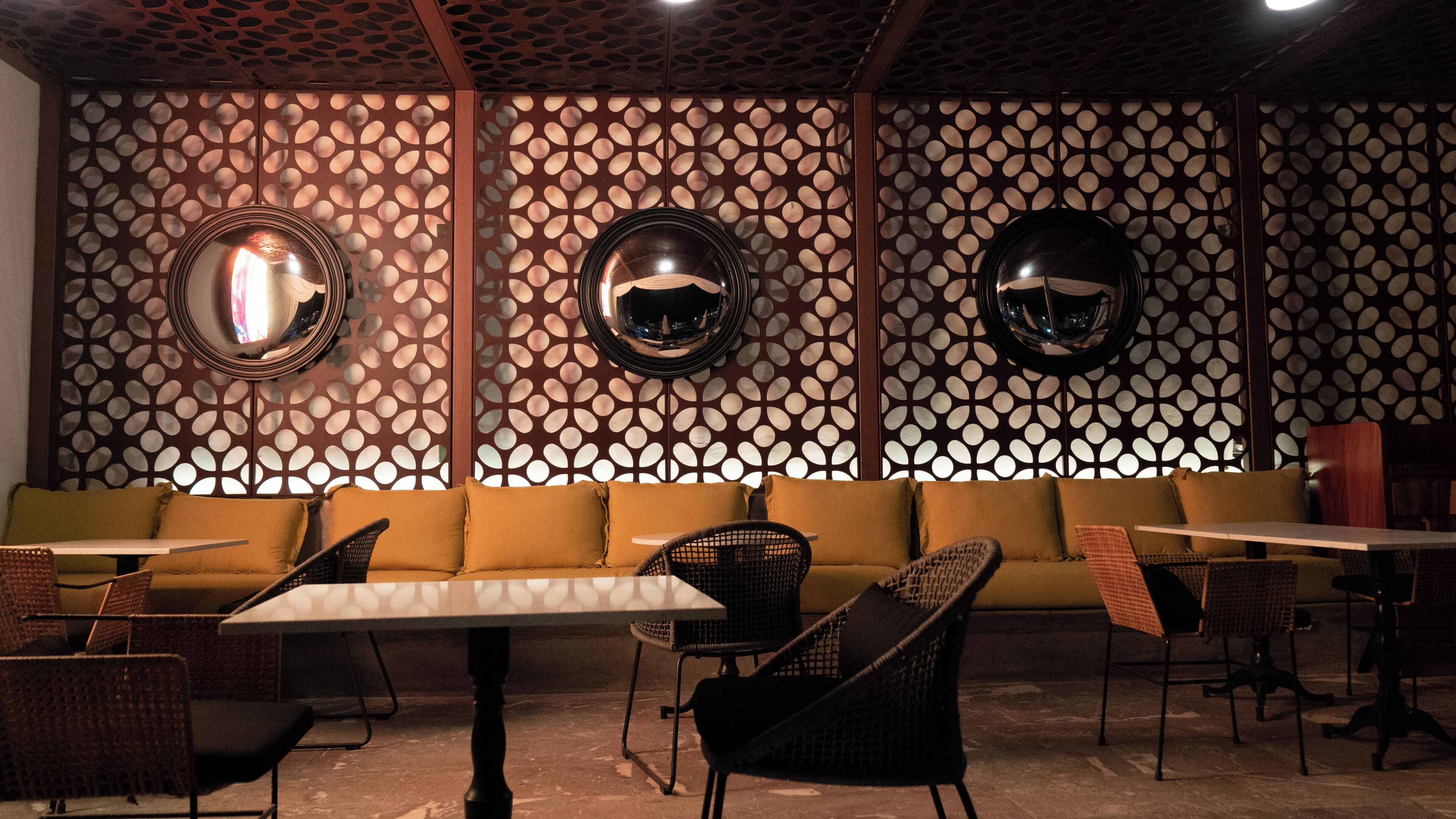 A restaurant interior with a patterned wall, round mirrors, a sofa, and wicker chairs.