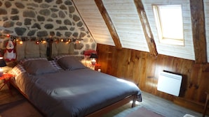 A cozy bedroom with a stone wall, wooden beams, a bed with gray bedding, a nightstand with a lamp, and a window.