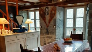 A rustic dining area with a wooden table, chairs, and a checkered tablecloth. There is a window with a view of the outside, a stone wall, and a wooden ceiling.