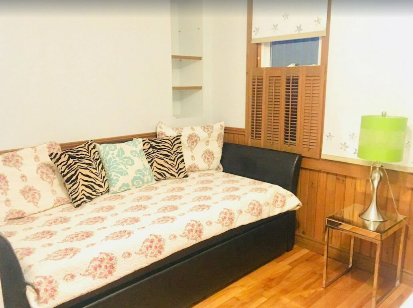 Een slaapkamer met een bed, nachtkastje, een raam met luiken en een groene lamp.
