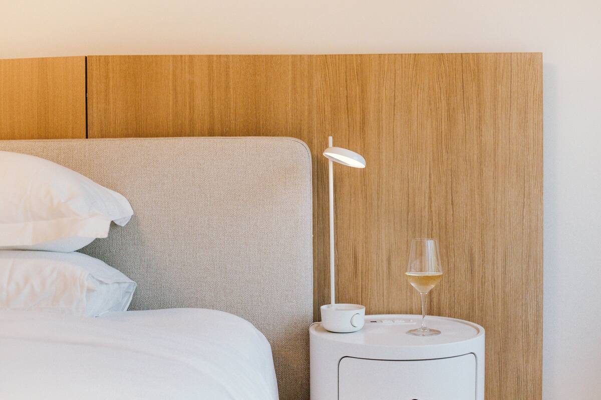 Ein ordentlich bezogenes Bett mit weißen Leinen und Kissen, ein Nachttisch mit einer weißen Lampe und einem Glas Weißwein sowie ein Holzkopfende.