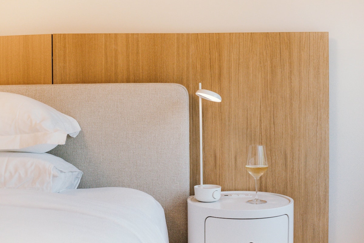 Una cama bien tendida con sábanas y almohadones blancos, una mesita de noche con una lámpara blanca y una copa de vino blanco, y un cabecero de madera.