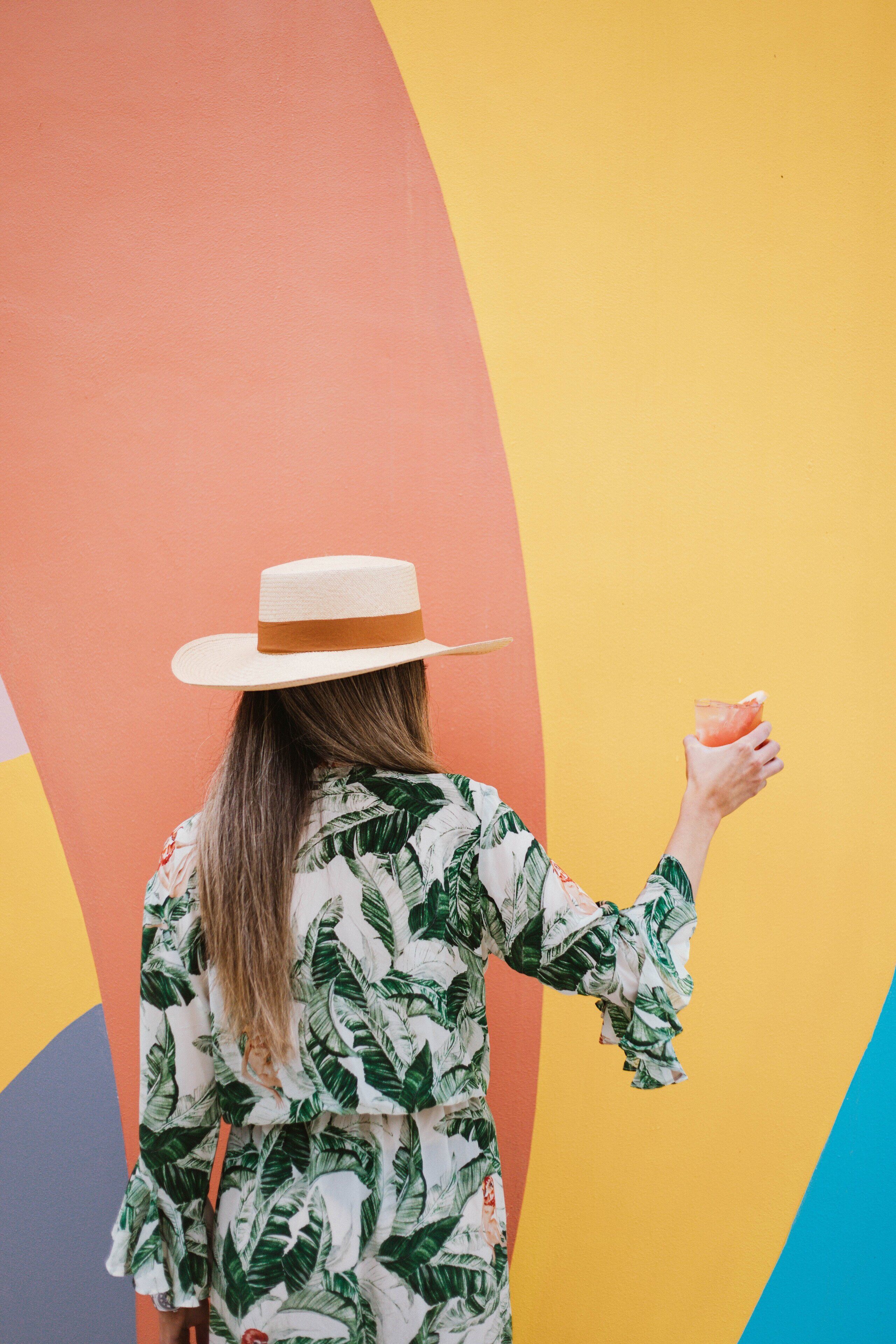 A person wearing a hat and a tropical print dress standing against a colorful wall.