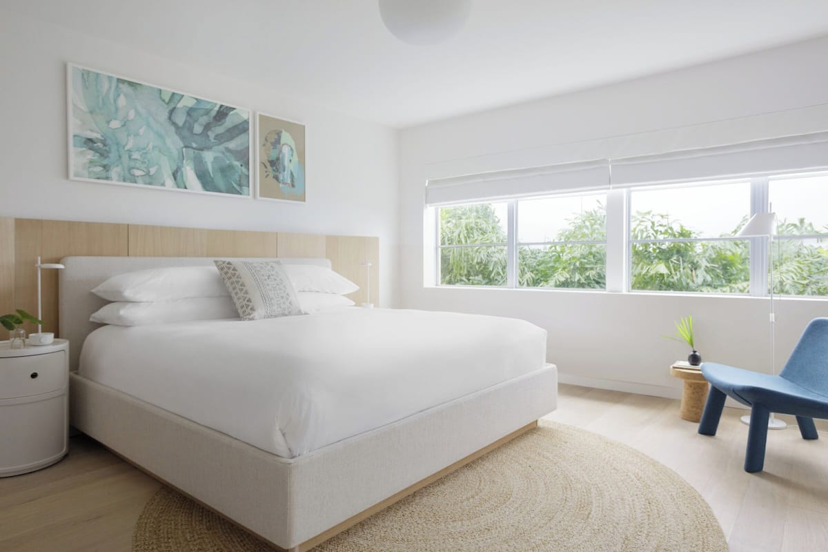Un dormitorio moderno con una cama grande, una mesita de noche, un sillón azul y una ventana con vistas a zonas verdes.