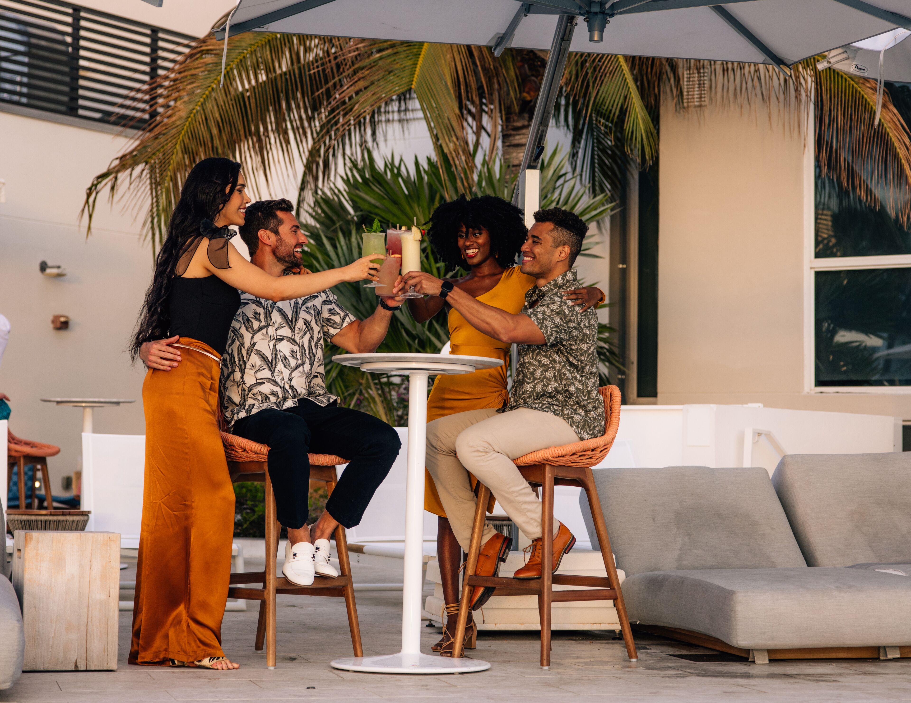 A group of people toasting around a table in an outdoor setting with a palm tree and a modern building in the background.