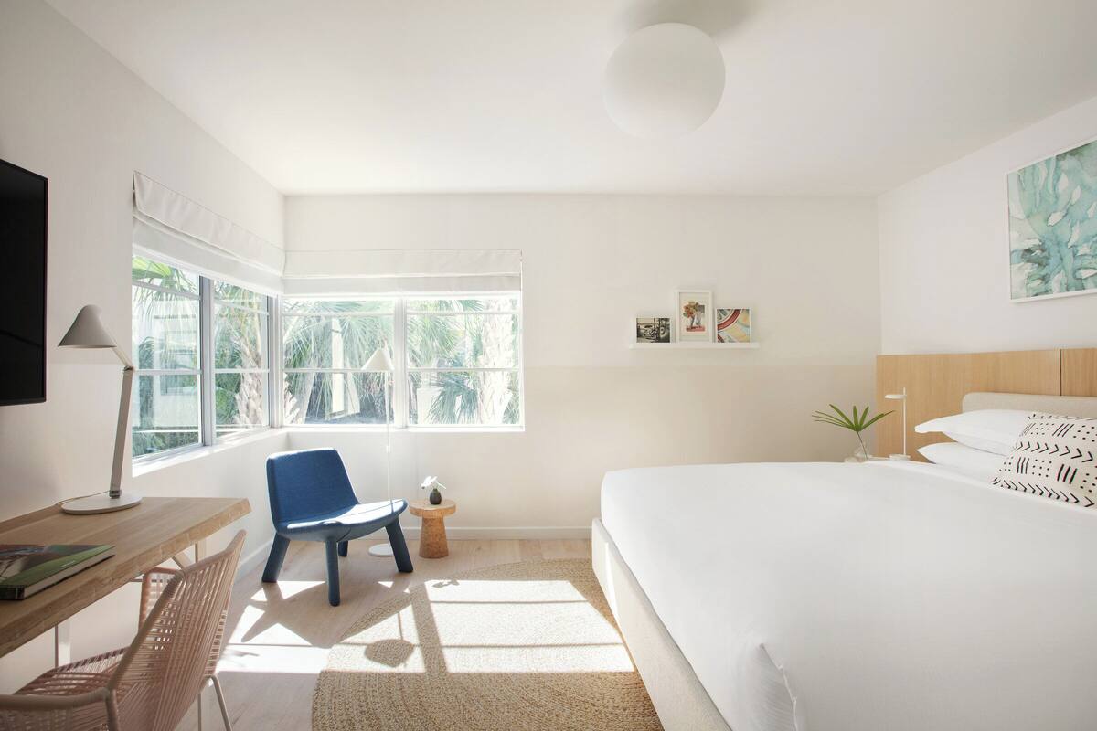 Ein helles, minimalistisches Schlafzimmer mit einem großen Bett, einem Schreibtisch, einem Stuhl und einem Fenster mit Blick auf Palmen.