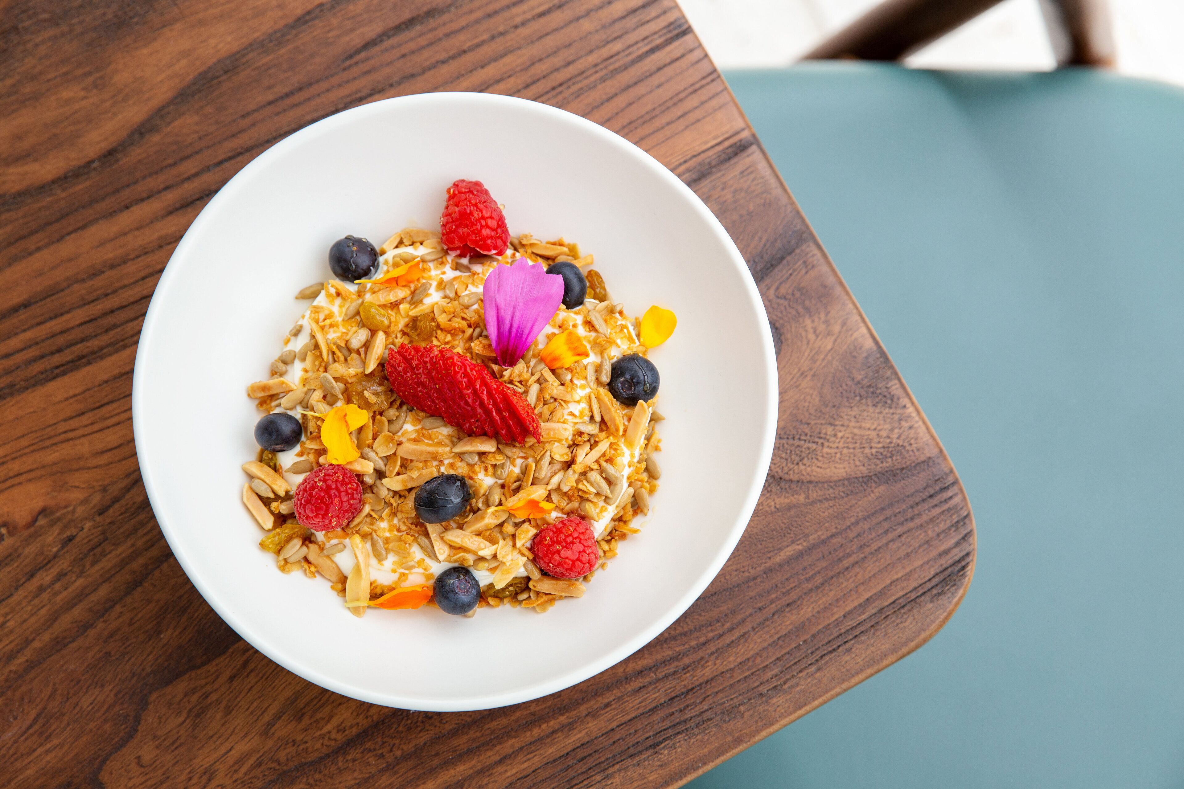 A bowl of cereal with fruits and a flower on a wooden table.
