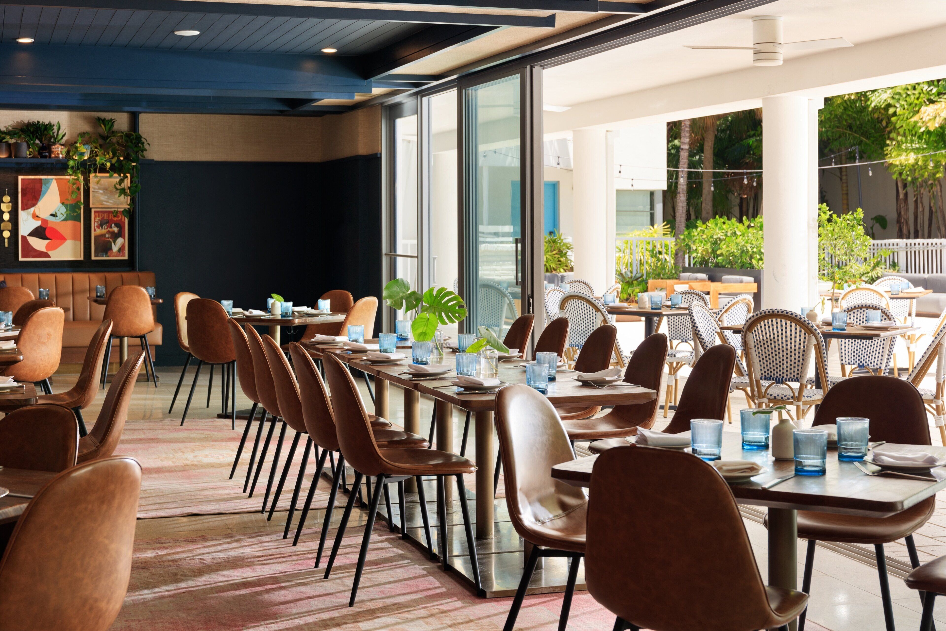 A modern dining area with multiple tables and chairs, large windows, and indoor plants.