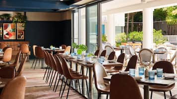 A modern dining area with multiple tables and chairs, large windows, and indoor plants.