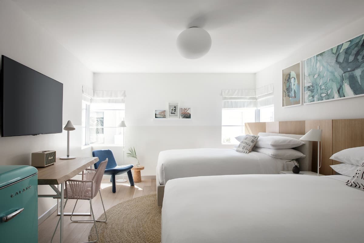 Quarto de hotel moderno com duas camas, uma escrivaninha, uma televisão e uma janela grande.