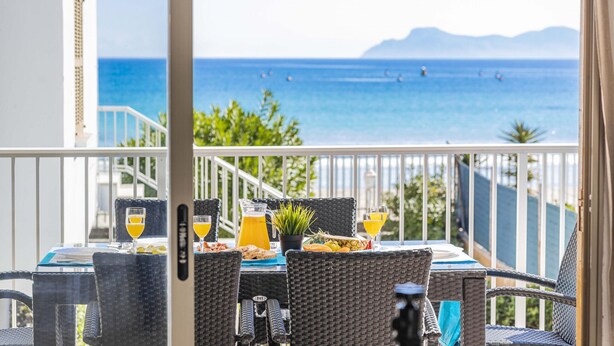 A balcony with wicker furniture, a table set with plates, glasses, and a pitcher of orange juice, overlooking a beach with boats and mountains in the distance.