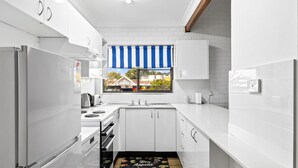 A modern kitchen with white cabinets, a stainless steel refrigerator, a stove, and a window with a blue and white striped awning. - Beach Street Bliss - situated in the heart of Woolgoolga (Woolgoolga)
