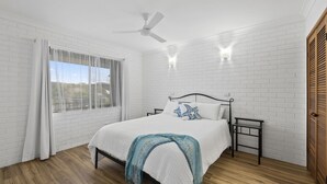 A bedroom with a bed, a ceiling fan, a window with curtains, and a brick wall. - Beach Street Bliss - situated in the heart of Woolgoolga (Woolgoolga)