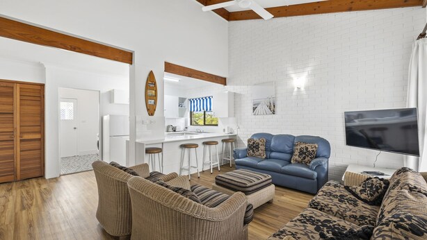 A spacious living area with a blue sofa, wicker chairs, a kitchen with white cabinets, and a flat-screen TV. - Beach Street Bliss - situated in the heart of Woolgoolga (Woolgoolga)