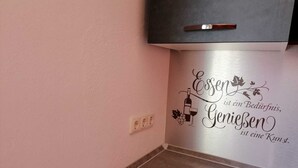A kitchen with a wall-mounted stainless steel range hood, a white countertop, and a decorative wall decal featuring a wine bottle and grapes. - Ferienwohnung 55 m², 1 Schlafzimmer, 1 Wohnzimmer, 1 Küche, 1 Bad, 1 Flur (Ensch)