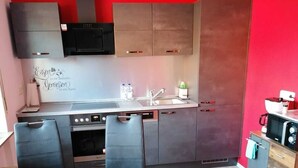 A compact kitchen with a red wall, a built-in oven, a microwave, and a sink. There are two black chairs and a small dining table. - Ferienwohnung 55 m², 1 Schlafzimmer, 1 Wohnzimmer, 1 Küche, 1 Bad, 1 Flur (Ensch)