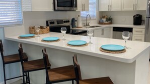 Una cocina moderna con armarios blancos, una isla central con taburetes, electrodomésticos de acero inoxidable y un amplio ventanal con persianas.