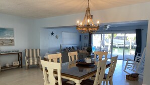 Un comedor con mesa de madera y sillas blancas, una araña y vistas al exterior a través de amplias ventanas.