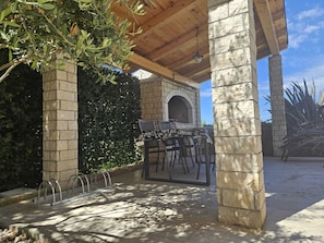 An outdoor patio with stone pillars, a covered seating area, and a stone wall with greenery.