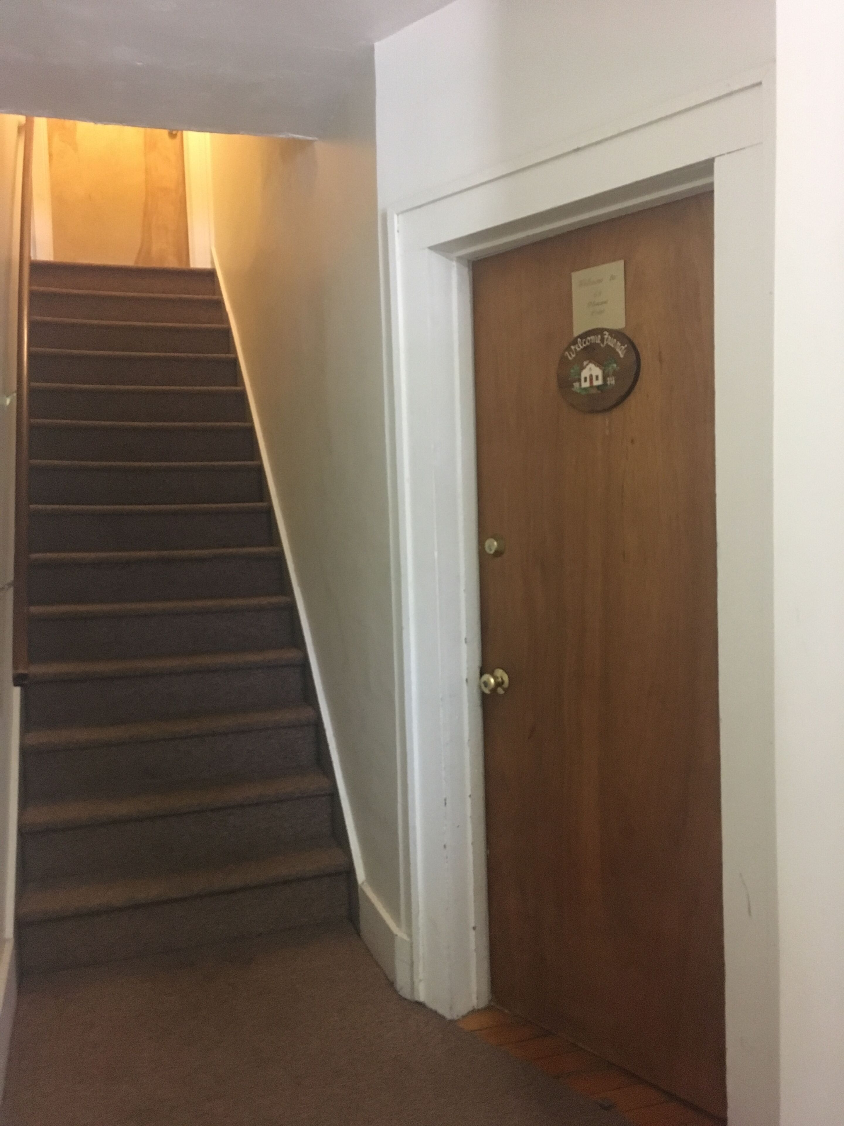 Una puerta de madera con una placa de bienvenida, una escalera alfombrada y una pared blanca.