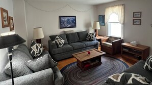Een woonkamer met een bank, fauteuils, een salontafel, een lamp, een vloerkleed en een raam met gordijnen.
