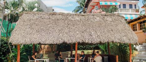 A thatched-roof outdoor seating area with tables and chairs, surrounded by greenery and a building with a balcony in the background.