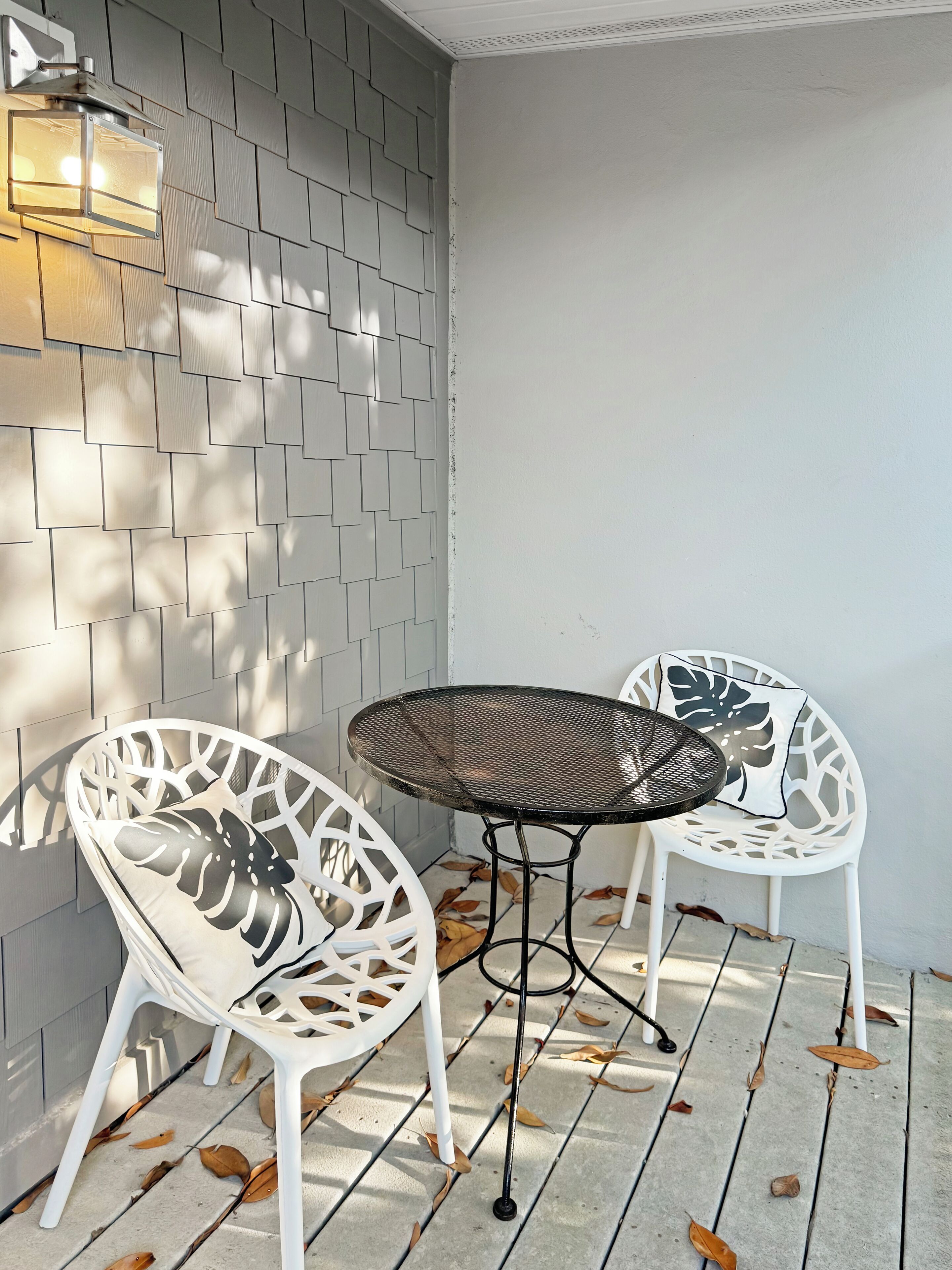 En patio med to hvide metalstole og et rundt bord, en vægmonteret lampe og et flisegulv med spredte blade.