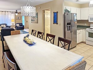 A dining area with a long table, chairs, a chandelier, and a kitchen with appliances. - STEPS FROM PRIVATE GATED BEACH, RENOVATED 4 BEDRM w/ 2 BALCONIES & 2 PATIOS (Miramar Beach)