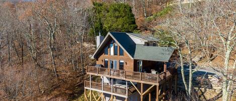 A wooden cabin with a balcony, surrounded by trees and a rocky hillside.
