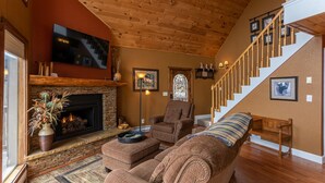 A cozy living room with a fireplace, a large TV, a staircase, and a comfortable seating area. - Grand Panorama - Pet friendly! Sunrise AND Sunset Views, Hot Tub (Banner Elk)