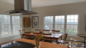 A dining area with a wooden table and chairs, a grill outside the window, and a large window with a view of trees. - Spectacular Views and Sunsets in the Lake Sunapee Region (New London)