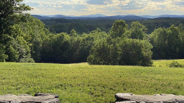 A stone bench overlooking a grassy field with a forested mountain range in the distance. - Spectacular Views and Sunsets in the Lake Sunapee Region (New London)
